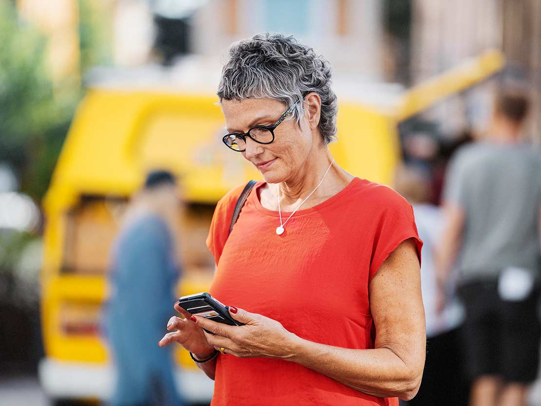 Äldre kvinna står utomhus och tittar på sin mobiltelefon