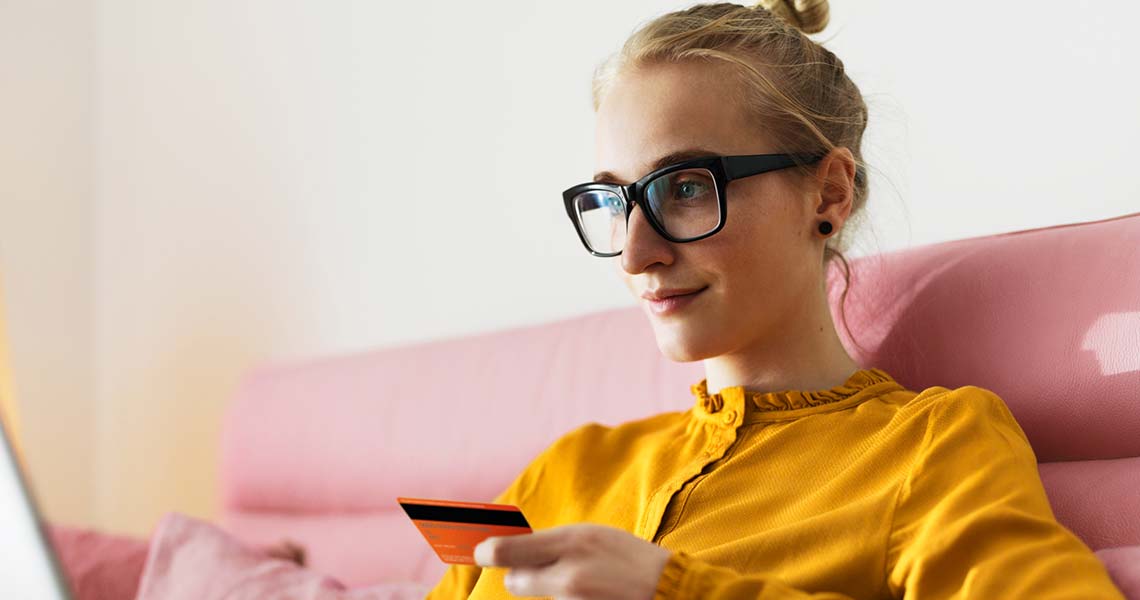 Tjej i glasögon och gul tröja som sitter i rosa soffa med betalkort och laptop