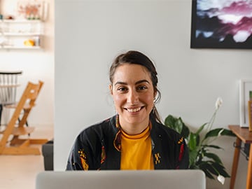 Ung tjej som sitter vid laptop ser leendes in i kameran